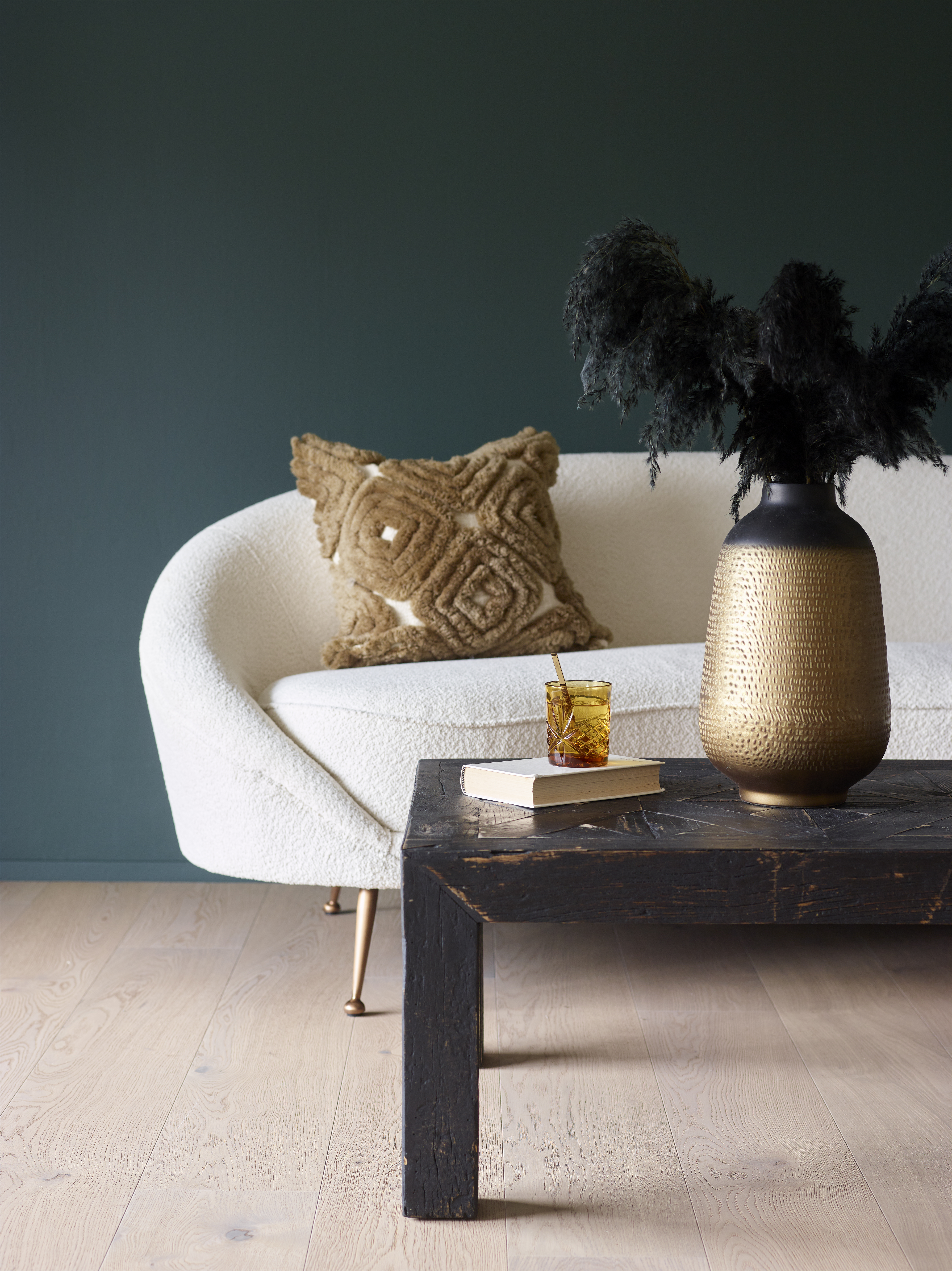 Kremhvit boucle-sofa med frynsepute mot petroleumsgrønn vegg, sofabord med bok, drink og gyllen vase med svart pampasgress.